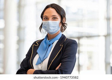 Portrait Of Flight Attendant Standing At Airport Terminal Wearing Face Mask For Safety Against Covid-19. Stewardess With Surgical Mask Looking At Camera, Aircrafts Are Waiting And Preparing Flight.