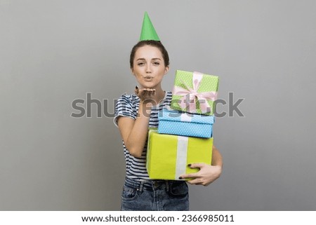 Portrait of flirting woman wearing striped T-shirt and party cone, holding stack of present boxes, sending air kiss. Indoor studio shot isolated on gray background.
