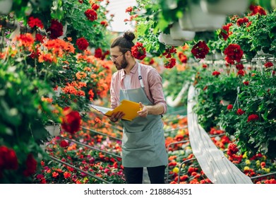 Portrait Of A Florist Man Holding Clipboard For A Checklist While Working In A Plant Nursery. Plantation Of Flowers In The Background. Gardening, Profession And People Concept. Wearing An Apron.