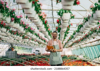 Portrait Of A Florist Man Holding Clipboard While Working In A Plant Nursery. Plantation Of Flowers In The Background. Gardening, Profession And People Concept. Looking Into The Camera. Copy Space.