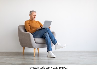 Portrait of focused mature man using laptop sitting on armchair. Confident male adult working on pc, typing on keyboard isolated on white studio background wall, full body length, free space mockup
