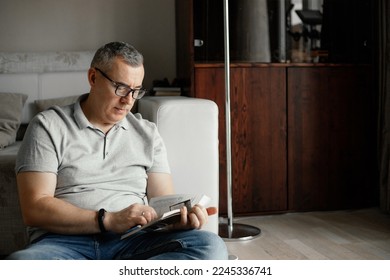 Portrait of focused middle-aged man with short hair wearing grey T-shirt, glasses, jeans, sitting near sofa on floor, holding reading book attentively at home. Spending free time, hobby, education. - Powered by Shutterstock