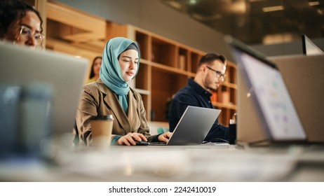 Portrait of Focused Muslim Woman Using Laptop in a Contemporary Spacious Office. Account Specialist Smiling While Having Productive Day. Active Colleagues Working on Computers in the Background. - Powered by Shutterstock