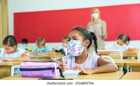 Portrait of focused schoolgirl wearing protective face mask working at lesson in classroom, writing exercise