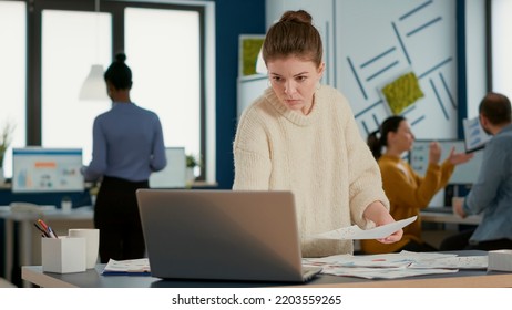 Portrait Of Focused Startup Employee Looking At Laptop And Charts Comparing Sales Statistics Standing At Desk. Woman In Busy Office Sorting Through Business Papers Looking At Data Analytics.