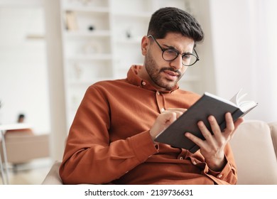 Portrait of focused young Arabic male trying to read paper book, squinting to see more clearly, wearing glasses, having difficulties seeing text because of vision problems, sitting on couch - Powered by Shutterstock