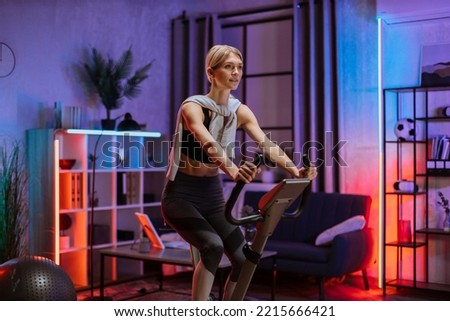 Portrait of focused young female wearing sport bra and using exercise bike. Home fitness workout sporty woman training on smart stationary bike indoors at evening time. Young caucasian girl athlete.