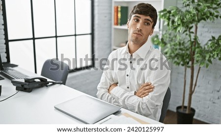Portrait of a focused young hispanic man, a serious business worker, brooding over work complexities at his office desk, arms crossed in a thinking gesture.