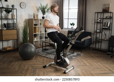 Portrait of focused young male with water bottle wearing sportswear using exercise bike. Home fitness workout sporty man training on smart stationary bike indoors. Young caucasian guy athlete. - Powered by Shutterstock