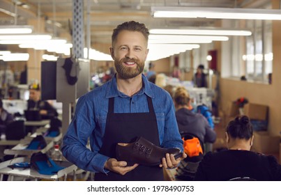 Portrait Of Footwear Factory Worker With Brand New Product. Happy Young Man Standing In Workshop, Holding New Leather Boots, Looking At Camera And Smiling. Shoe Manufacturing Industry Concept