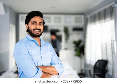 Portrait of formal business man confident successful. Indian businessman or manager in light blue shirt, stands at workplace in office, arms crossed, looks directly at camera and smiles friendly - Powered by Shutterstock