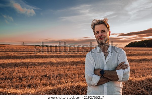 Portrait of forty years old, caucasian farmer in\
white shirt standing smiling in front of mowed wheat field\
(stubble). Agriculture - harvest time. Country outdoor scenery,\
warm sunset light.