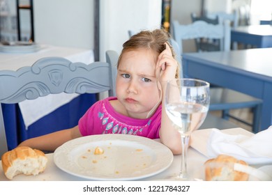 Portrait Of Four Years Old Blonde Little Girl With Pink Eating Bread, Looking At With Angry Expression Face, Sitting Indoor In Restaurant