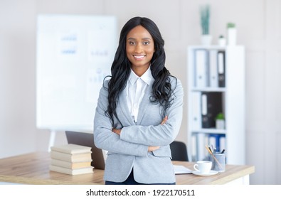 Portrait of friendly black female manager in formal suit standing with crossed arms at contemporary office. Successful African American busineswoman posing and smiling at camera