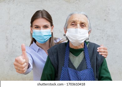 Portrait Of Friendly Caregiver Posing With Elderly Ill Woman Wearing Surgical Mask Because Of Covid-19 Pandemic, Showing Thumbs Up