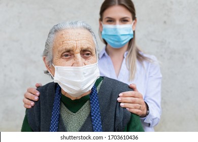 Portrait Of Friendly Caregiver Posing With Elderly Ill Woman Wearing Surgical Mask Because Of Covid-19 Pandemic