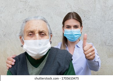 Portrait Of Friendly Caregiver Posing With Elderly Ill Woman Wearing Surgical Mask Because Of Covid-19 Pandemic, Showing Thumbs Up