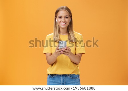 Portrait of friendly charismatic and outgoing good-looking young female in yellow t-shirt holding smartphone wearing wireless earphones enjoying listening music on way to fitness gym