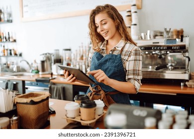 Portrait of a friendly female barista holding a tablet computer and smiling. A young coffee shop owner standing behind the bar using a digital tablet. Takeaway concept, technology. - Powered by Shutterstock