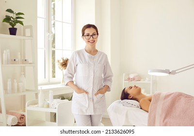 Portrait of friendly female woman beautician, aesthetic nurse or masseuse at her workplace. Smiling female beauty salon worker standing next to her client lying on couch in bright office. - Shutterstock ID 2133626095