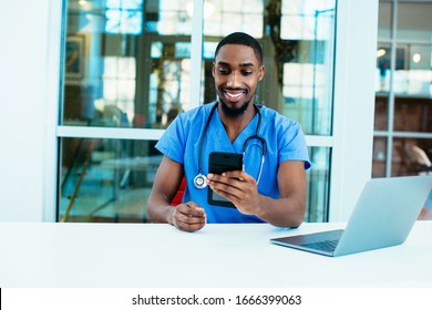 Portrait Of A Friendly Male Doctor Or Nurse Wearing Blue Scrubs Uniform And Stethoscope Sitting At Desk With Laptop In Hospital Checking Mobile Phone