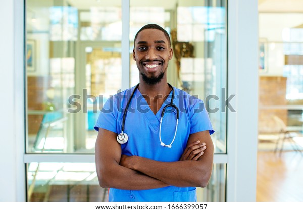 Portrait of a\
friendly male doctor or nurse wearing blue scrubs uniform and\
stethoscope, with arms crossed in\
hospital