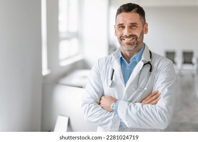 Portrait of friendly middle aged european male doctor in workwear with stethoscope on neck posing with folded arms in clinic interior, looking and smiling at camera, free space
