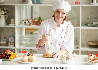 Portrait of friendly smiling female professional confectioner topping a cupcake with cream using a pastry bag. Looking at the camera. Indoors image. Pastry chef woman making creamy cakes - Powered by Shutterstock