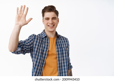 Portrait Of Friendly Smiling Man Waving Raised Hand, Saying Hello And Looks Happy, Greeting You Make Hi Wave Gesture, Welcome Friends, Stands Against White Background