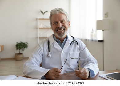 Portrait Of Friendly Smiling Senior Adult Healthcare Professional Therapist Sitting At Workplace. Happy Confident Older Male Doctor Physician Wearing White Medical Coat Stethoscope Looking At Camera.