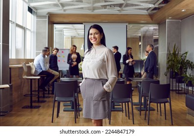 Portrait of friendly and successful business woman standing against background of colleagues in office. Millennial female leader or coach stands against background of people talking to each other. - Shutterstock ID 2244847179