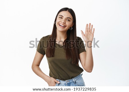 Portrait of friendly young happy woman waiving hand to say hi, greeting you with hello gesture, saying goodbye, standing over white background