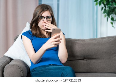 Portrait of frighten shocked girl, scared young stressed anxious woman is looking at her cell mobile phone, reading negative news, message at smartphone, sitting at couch, sofa at home in living room