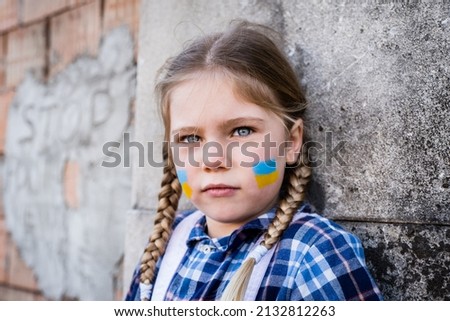 Portrait of a frightened child girl with painted flag on the cheek in yellow-blue colors of the Ukrainian flag. Peace and protection children concept