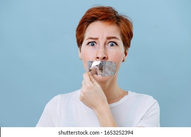 Portrait Of A Frightened Middle Aged Woman With Short Ginger Hair And Her Mouth Scotch Taped. Tearing Tape Off Of Her Mouth. She's Wearing White T-shirt. Over Blue Background.