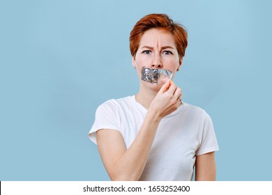 Portrait Of A Frightened Middle Aged Woman With Short Ginger Hair And Her Mouth Scotch Taped. Tearing Tape Off Of Her Mouth. She's Wearing White T-shirt. Over Blue Background.