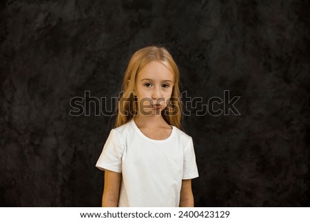 Portrait of frowning cover girl kid model 6 year old in white t-shirt expression emotion, angry looking at camera. Scowl child posing at black, studio shot. Kids emotional concept. Copy ad text space