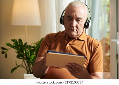 Portrait Of Frowning Senior Man Wearing Headphones When Watching Popular Science Video On Tablet Computer