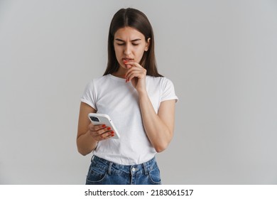 Portrait of a frowning young casual white woman in t-shirt with long brunette hair standing over gray wall background holding mobile phone - Shutterstock ID 2183061517