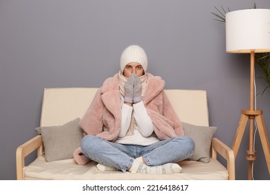 Portrait of frozen beautiful woman wearing warm clothes and wrapped in scarf sitting on sofa in room, trying to get warm in cold apartment, looking at camera. - Shutterstock ID 2216418617