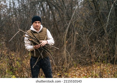 Portrait Of Frozen Tourist Male Wearing Warm Clothes Collecting Dry Deadwood For Fire In Forest On Overcast Cold Day. Concept Of Scout, Research, Travel And Survival In Nature.