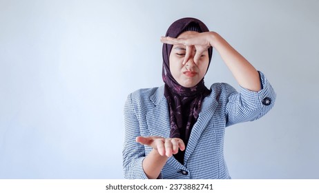 Portrait of frustrated Asian woman wearing hijab and formal wear pinching nose with disgust on his face due to bad smell isolated over gray background.