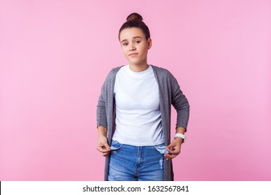 Portrait Of Frustrated Brunette Teenage Girl With Bun Hairstyle In Casual Clothes Showing Empty Pockets Inside Out, Poor Has No Money, Allowance. Indoor Studio Shot Isolated On Pink Background