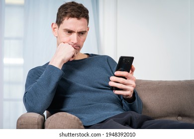 Portrait of frustrated confused frown guy, young sad upset handsome man is looking at screen his cell mobile phone, get negative news on smartphone, sitting at home on sofa or couch in living room