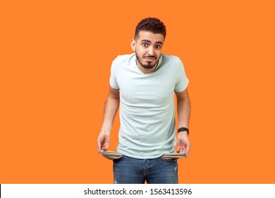 Portrait Of Frustrated Worried Brunette Man With Beard In Casual White T-shirt Turning Out Empty Pockets Showing I Have No Money Gesture, Bankrupt. Indoor Studio Shot Isolated On Orange Background