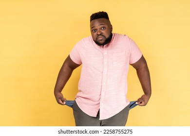 Portrait of frustrated worried man wearing pink shirt turning out empty pockets showing no money gesture, frowning face. Indoor studio shot isolated on yellow background.