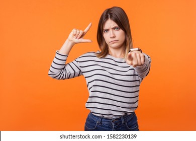 Portrait Of Frustrated Young Woman With Brown Hair In Long Sleeve Shirt Showing Looser Gesture And Pointing At Camera, Looking With Grumpy Face. Indoor Studio Shot Isolated On Orange Background