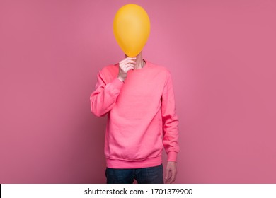 Portrait Full Lenght Of Man In Pink Sweater Cover Face Behind Yellow Balloon. Tall Incognito Male Person Stand Alone. Isolated Over Pink Background