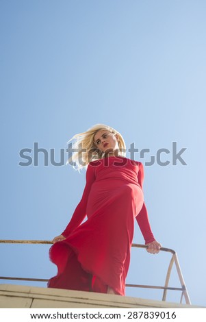 Portrait of full length body Woman in airy long red dress stand up and keep metal fence Blond hair fly in air Cute blondy girl against blue summer sky background Empty space for inscription 