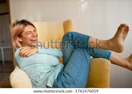 Portrait of funny adorable barefoot young female with pinkish hair and facial piercing laughing out loud having fun at home, sitting comfortably in armchair. Pretty girl relaxing indoors, smiling